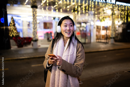 Young relaxed lady texting and wearing headphones with christmas lights behind. Happy girl listening to music at the street during winter using her phone.