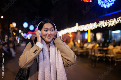 Young cheerful woman wearing headphones with christmas lights behind. Smiling girl listening to music at the street during winter.