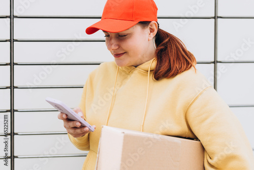 Young woman in red hat picks up parcel from automatic post office machine, Courier standing with phone and small box. Concept of fast delivery to automatic self lockers photo