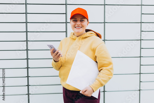 Young woman in red hat picks up parcel from automatic post office machine, Courier standing with phone and small box. Concept of fast delivery to automatic self lockers photo