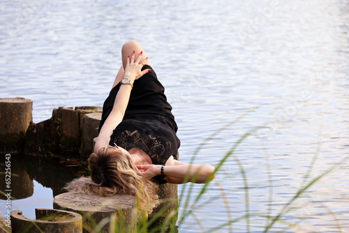 Woman in black dress lying on wooden logs on lake beach. Leisure in autumn park