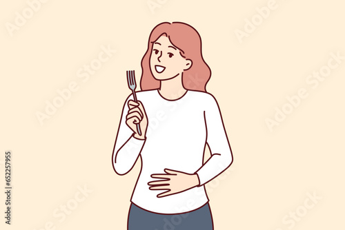 Hungry woman with fork in hands touches stomach, demonstrating good appetite and desire to dine. Hungry girl needs food and looks at screen with smile, waiting for waiter with order