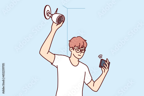 Man installs surveillance camera on wall and holds mobile phone, watching video through WIFI network. Guy establishes video surveillance system, wanting to secure home or office from criminals.