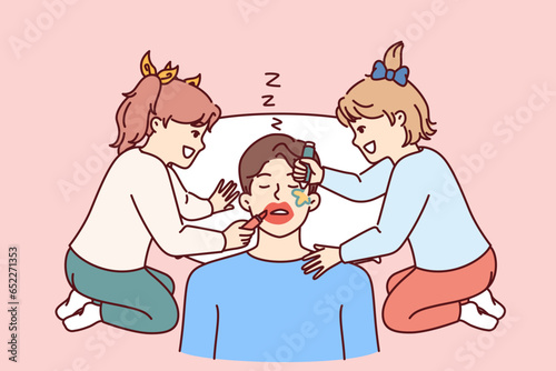 Funny little girls make up sleeping father by painting on man face with lipstick and colored markers. Two cheerful preteen daughters play pranks and laugh father sleeps after work.
