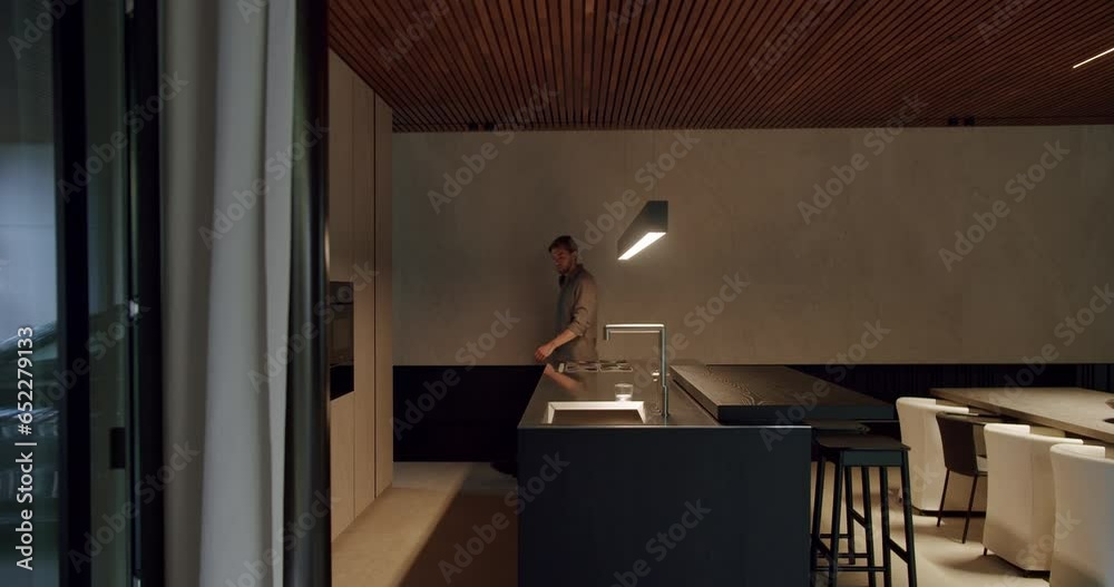 Young man goes to kitchen island with sink, take glass and fill drink water from faucet at modern apartment with stylish furniture and interior.