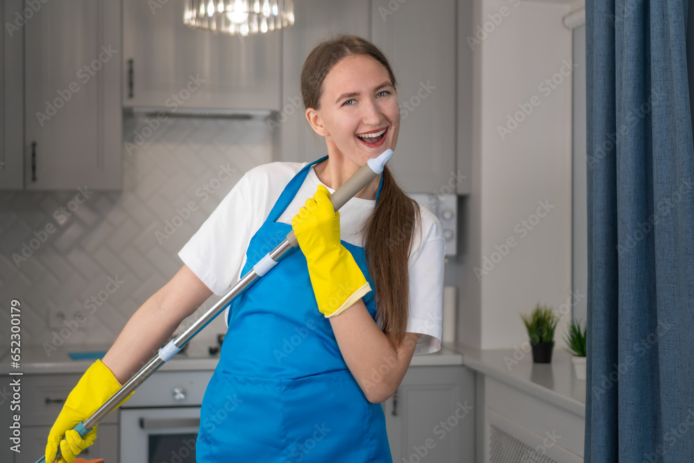 Happy young professional woman from a cleaning company cleans the house, sings into a mop as into a microphone, has fun.Housewife in an apron enjoys household chores, creatively engaged in cleaning