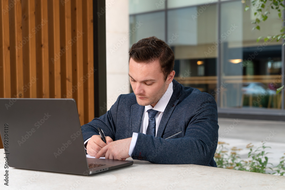 Young businessman in a suit and tie working on a laptop in a street cafe near the business center and making notes in a notepad. Man working on laptop with blank back panel without stickers Young businessman in a suit and tie working on a laptop in a street cafe near the business center and making notes in a notepad. Man working on laptop with blank back panel without stickers