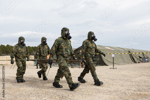 Vista de unos soldados vestidos con trajes de protección NBQ y mascaras antigás llevando una camilla.