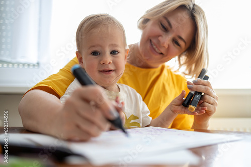 Middle aged blond mother drawing with colored pencils at the table at home with her little baby son 