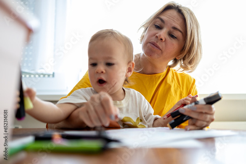 Middle aged blond mother drawing with colored pencils at the table at home with her little baby son 