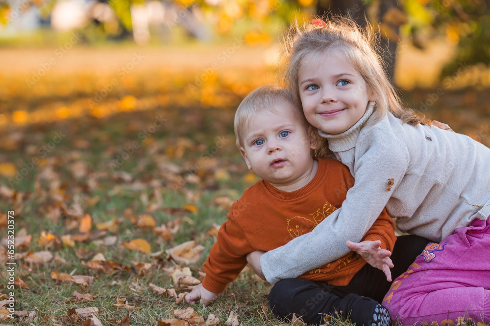 Portrait of a little boy 9 months old and a girl 4 years old outdoors. Happy children are playing in the autumn park. Sister hugs brother. Happy childhood and motherhood. Portrait of a little boy 9 months old and a girl 4 years old outdoors. Happy children are playing in the autumn park. Sister hugs brother. Happy childhood and motherhood.