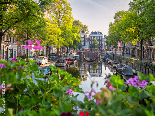 This photo showcases a peaceful Amsterdam canal scene. It includes an arched bridge, trees, boats docked in the canal and canal houses. Taken on a calm Sunday morning, the image reflects the canal's s photo