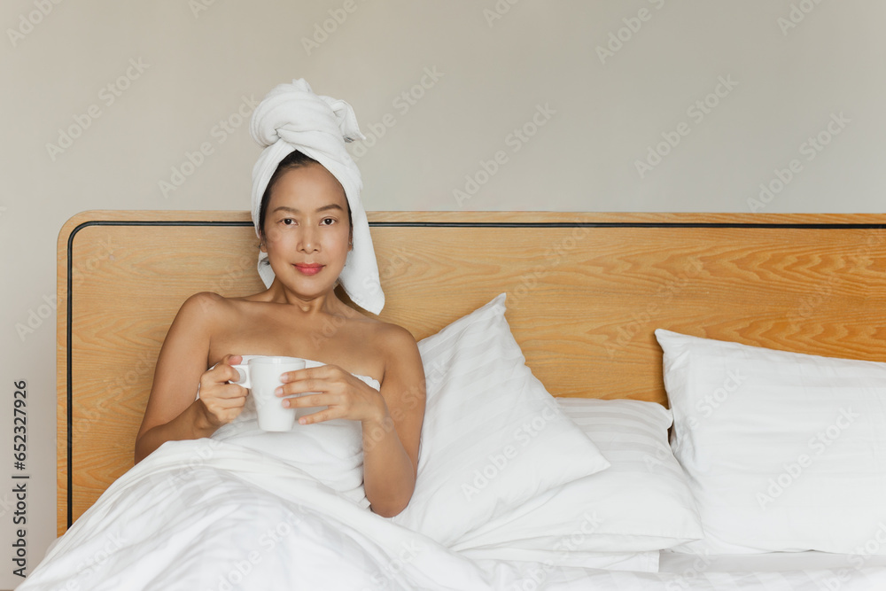 Woman lying in bed holding coffee mug with a towel on her head. Woman lying in bed holding coffee mug with a towel on her head.