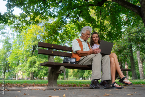 The grandfather and granddaughter are looking at photos on notebook, sitting on a bench in a city park. photo