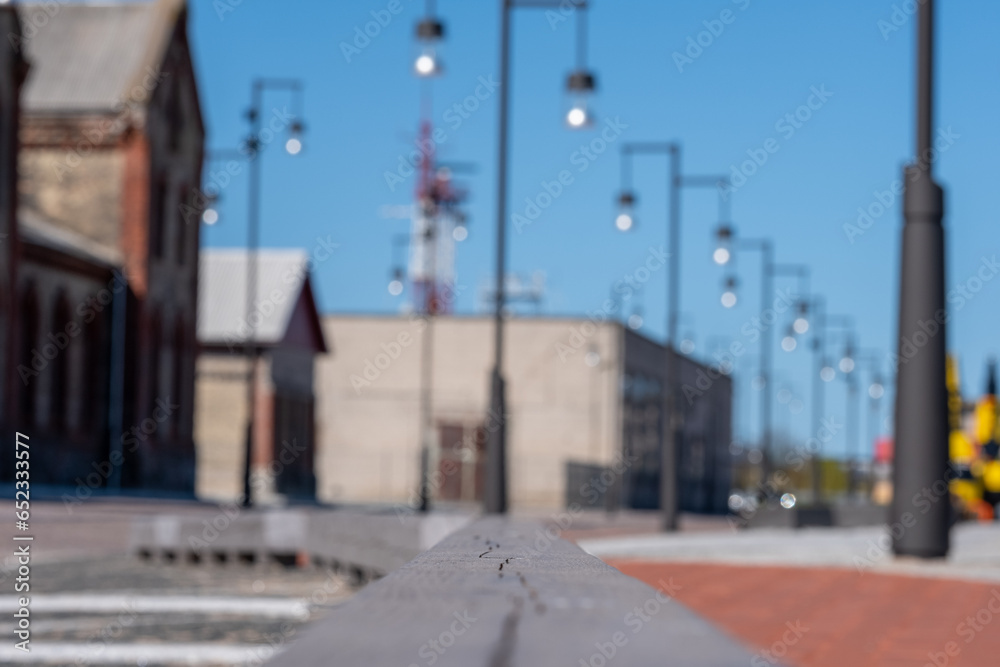 Industrial buildings, outdoor lanterns and a clear blue sky in blurred background. In the foreground, a wooden fence and a colorful pavement. Copyspace