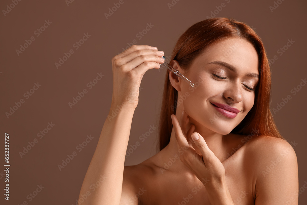 Fototapeta premium Beautiful young woman applying cosmetic serum onto her face on brown background, space for text
