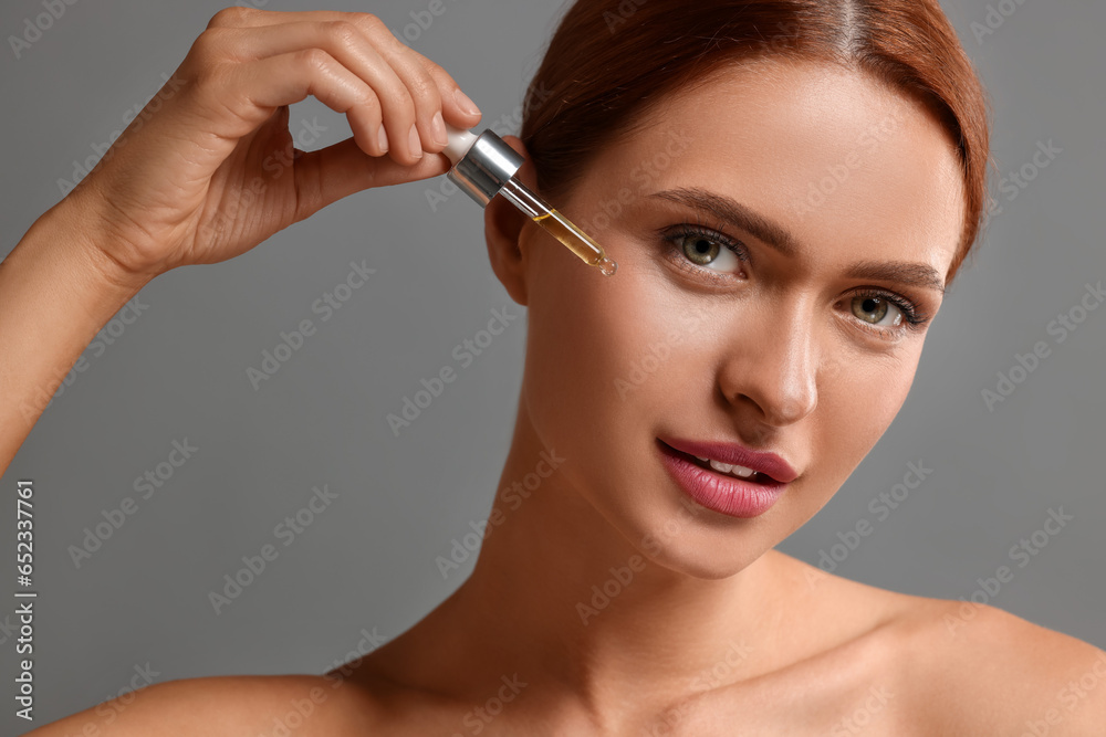 Fototapeta premium Beautiful young woman applying cosmetic serum onto her face on grey background, closeup