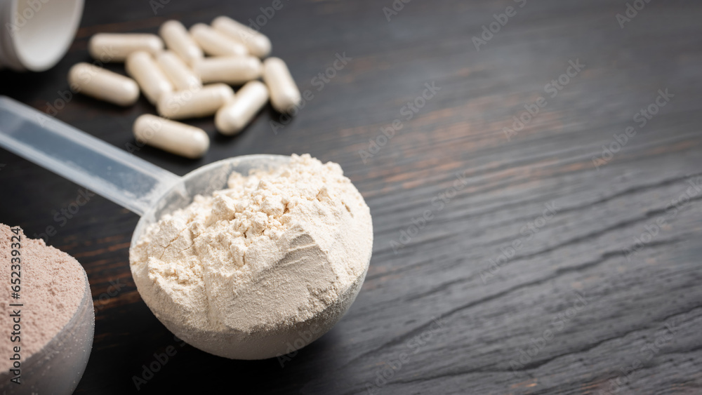 Scoop of whey or soy protein powder, white capsules of amino acids, vitamins and creatine, bodybuilding food supplements, sports nutrition on a dark wooden board