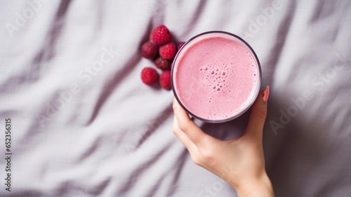 Close up of a hand holding a glass of raspberry smoothie on bed, top view angle