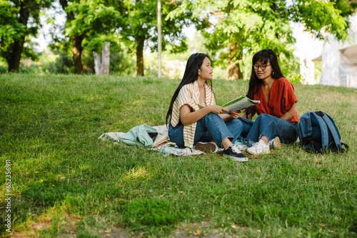 Fototapeta Naklejka Na Ścianę i Meble -  Two asian female students doing homework together while sitting on grass in park