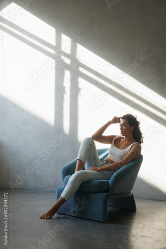 Beautiful brunette woman in white top and blue jeans sitting on blue armchair. Girl smile, happy. Portrait of young pretty woman. Smile and freshness. White background. Light from window, shadows