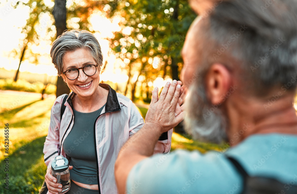 Fototapeta premium Sincere intelligent modern beautiful smiling cheerful active stylish older lady in sportswear giving high five to friend after morning workout outdoors. Senior couple jogging together.