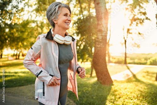 Remote technology and sport. Progressive aged lady with stylish grey haircut running in park with wireless headphones on neck. Slender female in sport outfit feeling contentment from morning jogging.