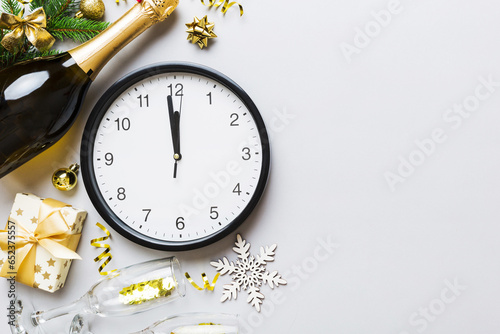 New year celebration concept with a bottle of champagne and two glasses toasting. Christmas gift box, alarm clock and fir tree branch on colored table. Top view Copy space