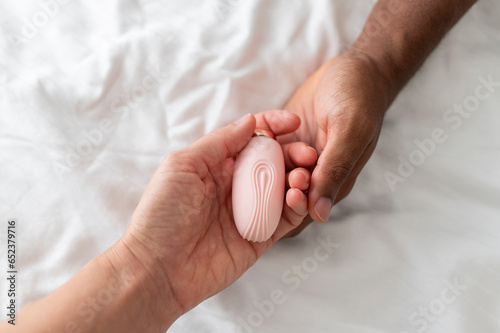 Pink sex toy vibrator for mutual pleasure in hands of white woman and African man, on the white bedding