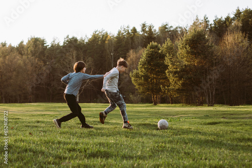 Foxy teenager grabbing friend on clothes attempting to overtake boy running towards ball, playing football in nature.