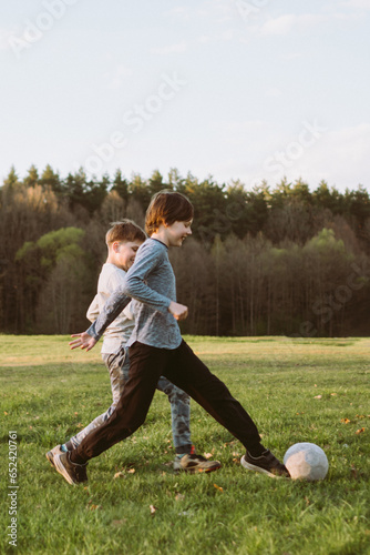 Joyful teen kicking ball while friend running close, active players practicing on lawn with happy expressions on faces.