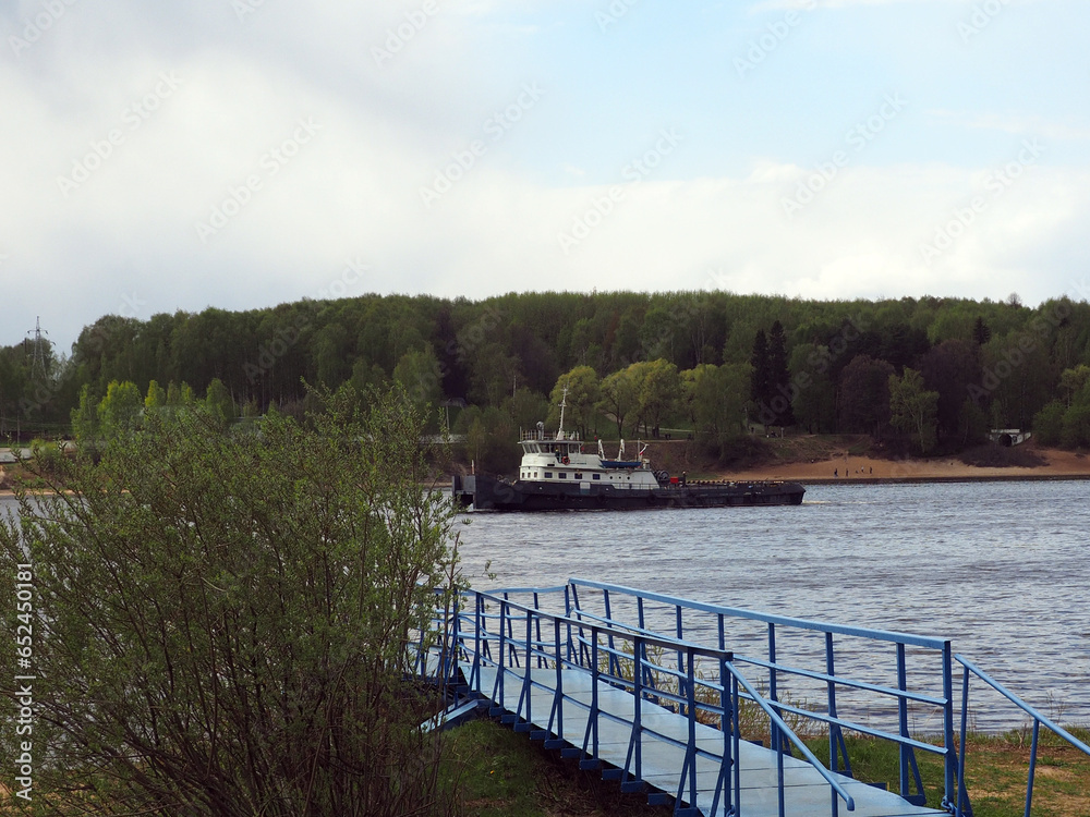The river tug o floats on the river against the background of the shore overgrown with trees and shrubs, leaving behind traces of foam. River and sea transportation, water transport.