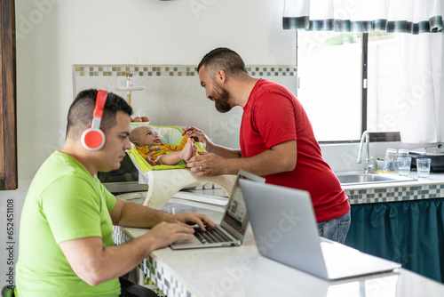 Image of a gay man working on his laptop with headphones in the dining room of their home and her husband tending to their adopted son in a baby chair on the concrete kitchen table.