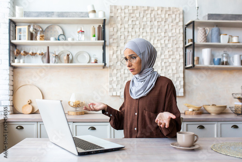Upset young arab woman in hijab sitting in front of laptop at home in kitchen and throwing up hands in frustration, talking on video call, received and reading bad news.