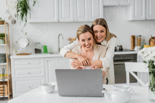 attractive blonde sisters looking at laptop smiling and hugging from behind, family bonding