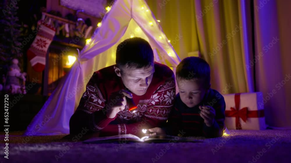 Young father with his baby boy reading book with flashlights lying in canvas tent on magical Christmas eve at cozy fireplace burning woods on background. Two generations family spending time together Young father with his baby boy reading book with flashlights lying in canvas tent on magical Christmas eve at cozy fireplace burning woods on background. Two generations family spending time together