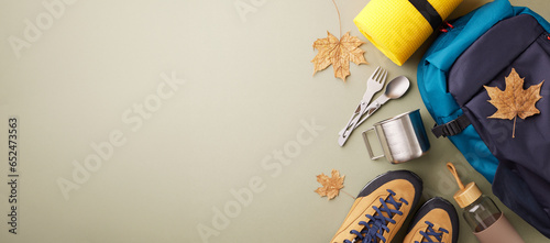 Weekend excursion into the natural world. Top view photo of metal utensils, hiking boots, backpack, trekking sticks, karemat, water bottle, autumn leaves on green background with advert zone