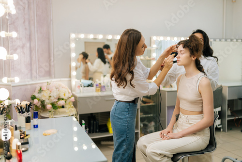 Wide shot of female hairdresser using hair tongs curling hair of young woman in beauty studio. Makeup artist applying foundation on lady face using sponge. Concept of make up and styling in 4 hands.