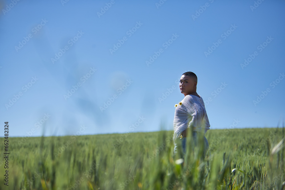 Young, South American, non-binary person, heavily makeup, posing in a white sweater with natural daisies, in the middle of a green wheat field. Concept queen, lgbtq+, pride, queer.