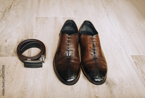 Men's brown leather stylish shoes and a black belt lie on the floor. Close-up photo, top view.