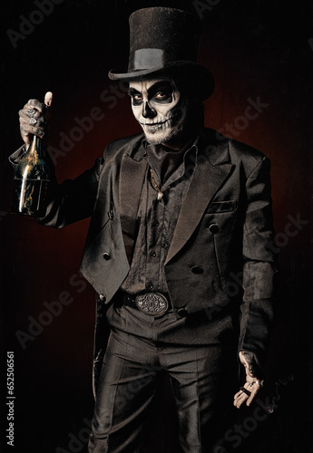 Young man in image of Baron Samedi, the Voodoo deity. Baron Saturday in black suit and top hat with a bottle of alcohol in hand. Day of the Dead (and Halloween) theme