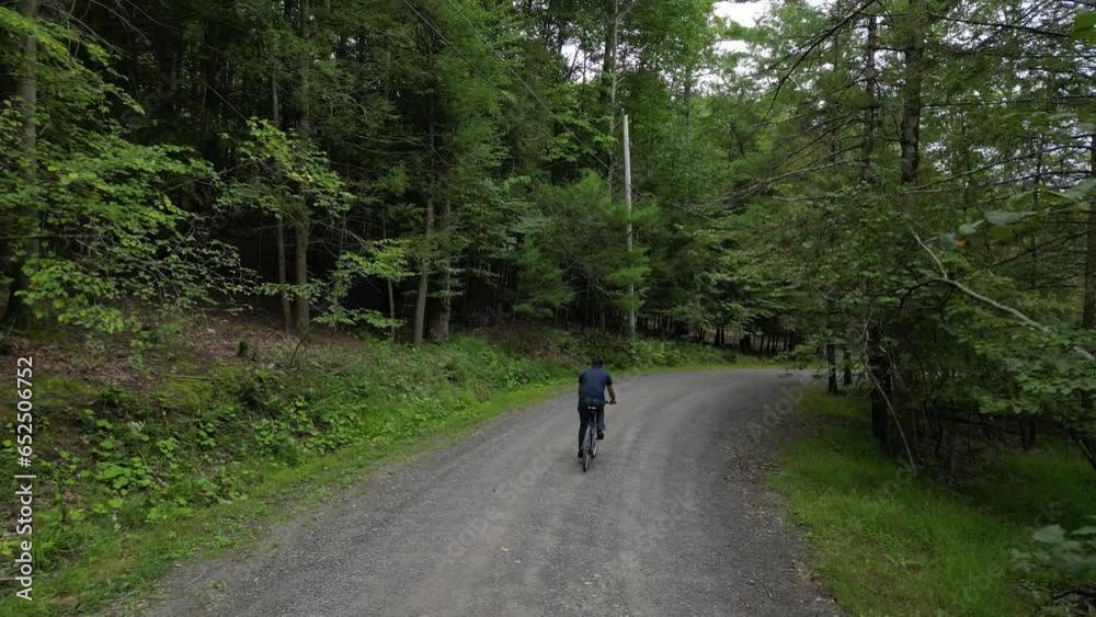 man riding bicycle down hill filmed from behind (footage from back, casual clothes, mountain bike, gravel path) descent, aerial footage, following descending rural road with trees man riding bicycle down hill filmed from behind (footage from back, casual clothes, mountain bike, gravel path) descent, aerial footage, following descending rural road with trees