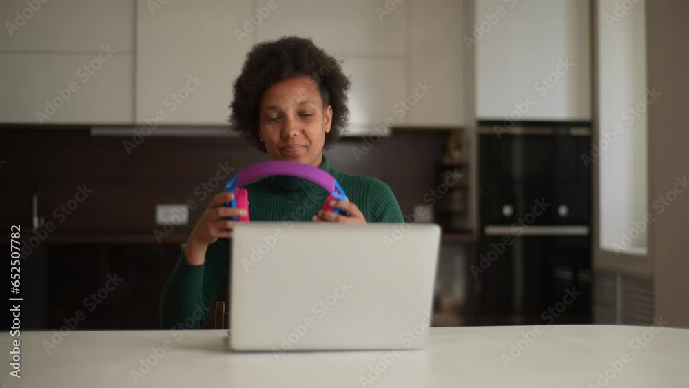 Portrait of happy smiling black female freelancer put on colorful wireless headphones. Front view of cheerful young woman using laptop at home listening music. Shooting in slow motion. Portrait of happy smiling black female freelancer put on colorful wireless headphones. Front view of cheerful young woman using laptop at home listening music. Shooting in slow motion.