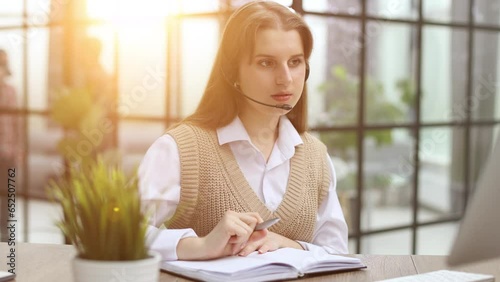 Attractive confident business woman, office manager, wearing headset using laptop while making, writing financial repor photo