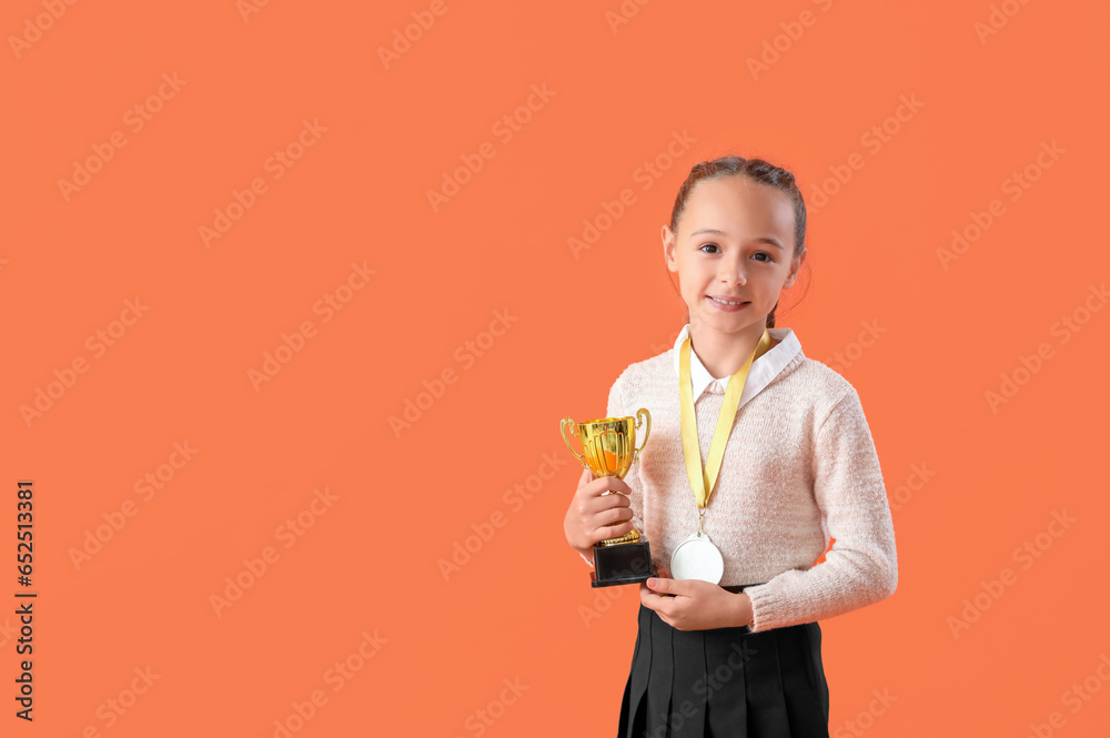 Smart little schoolgirl in stylish uniform with prize cup and medal on orange background Smart little schoolgirl in stylish uniform with prize cup and medal on orange background