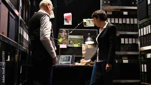 Law agents review police archive records in incident room, gathering intelligence from witness statements. Detectives team evaluating evidence authenticity, inspecting forensic report. photo