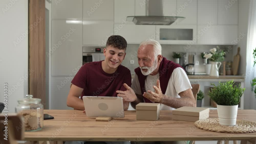 senior man grandfather pensioner gray hair and beard with his grandson teenager boy receive presents in box open read card happy smile in front of digital tablet at home online video call slow motion senior man grandfather pensioner gray hair and beard with his grandson teenager boy receive presents in box open read card happy smile in front of digital tablet at home online video call slow motion