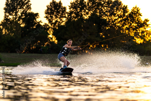 Female wakeboarder wakesurfing on sunset. Girl riding waterski cabel. Holding tow rope. Summer activities in the lake.