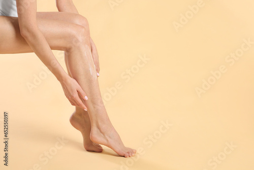 Woman applying body cream onto her smooth legs on beige background, closeup. Space for text