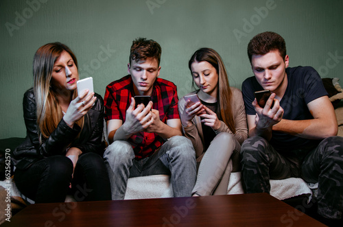 Group of four young adults male and female acting anti-social sitting in room and only looking at smartphones 